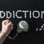 White pills spell out ADDICTION on a black background above a handcuffed fist attached to a pill bottle, representing prescription abuse challenges treated at Drug Rehab Fort Wayne Indiana.