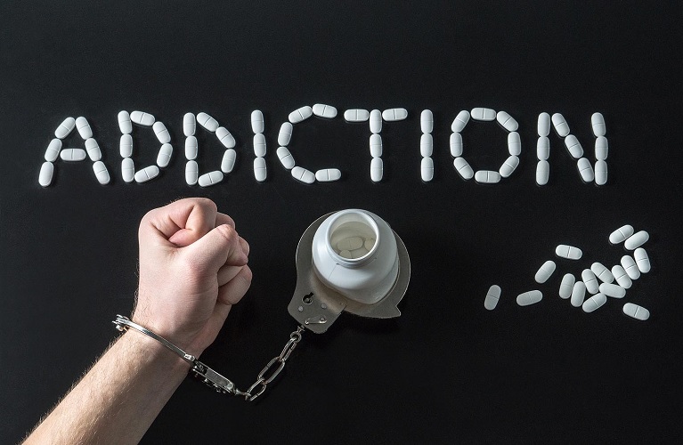 White pills spell out ADDICTION on a black background above a handcuffed fist attached to a pill bottle, representing prescription abuse challenges treated at Drug Rehab Fort Wayne Indiana.