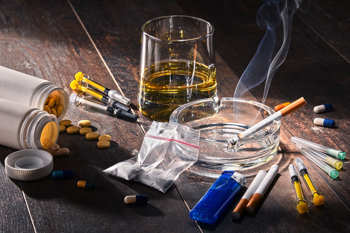 Various substances and drug paraphernalia, including alcohol, pills, syringes, cigarettes, and powder arranged on a dark wooden table.