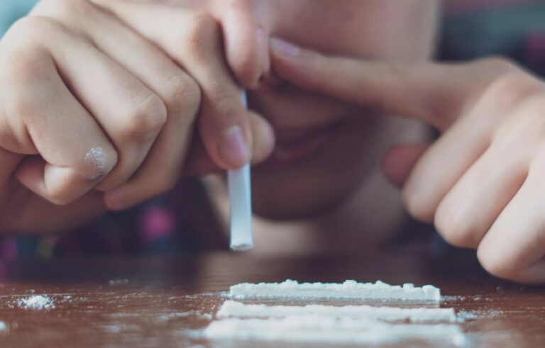 Close-up of a person using a tube to snort white powder lines off a table, showing actions causing long-term Cocaine Nasal Damage.