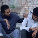 Young man placing a comforting hand on his friend's shoulder outdoors, illustrating support in enabling vs helping addiction.