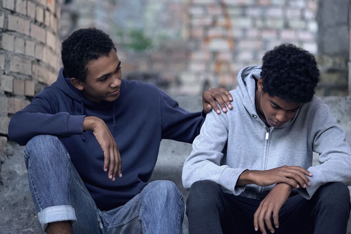 Young man placing a comforting hand on his friend's shoulder outdoors, illustrating support in enabling vs helping addiction.
