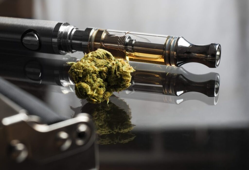 A sleek vape pen with an oil cartridge resting next to a dry green flower bud, reflecting cannabis vaping trends and devices on a dark surface.