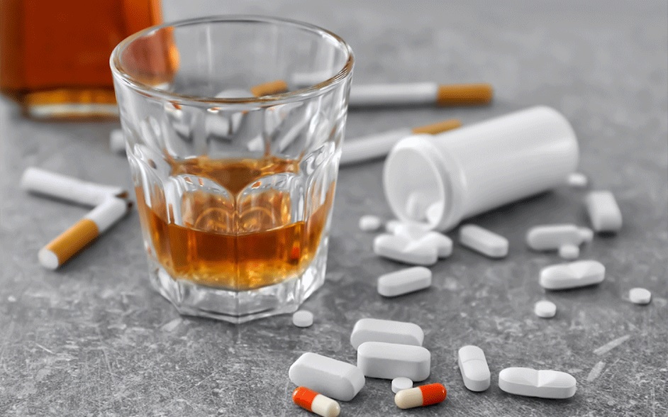 A glass of liquor surrounded by spilled white pills and cigarettes on a grey surface, representing addiction struggles solved by drug and alcohol rehab in Indianapolis.