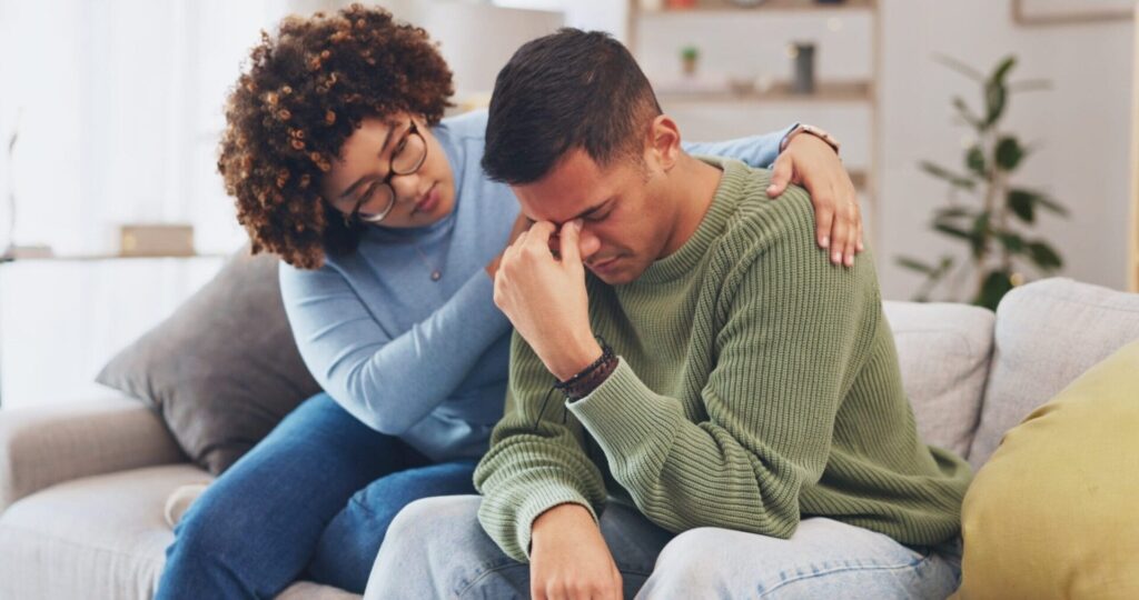 A supportive friend comforts a grieving man on a couch, illustrating the importance of emotional support when maintaining sobriety after loss.