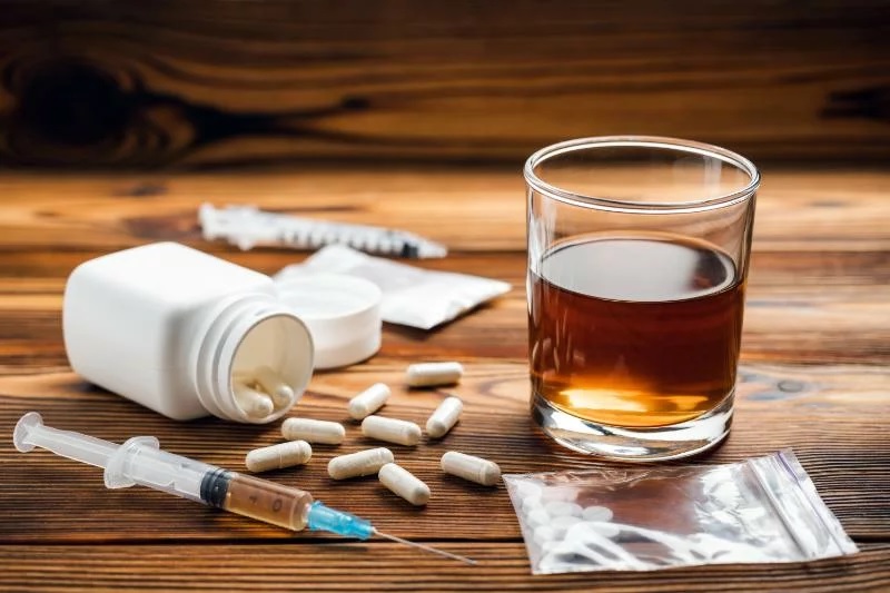 A glass of alcohol, syringes, and pills on a wooden surface, illustrating typical triggers encountered when navigating substance use treatment.