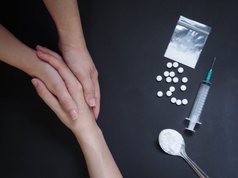 A supportive hand offer comfort beside pills and a syringe in a Prescription Drug Rehab setting.