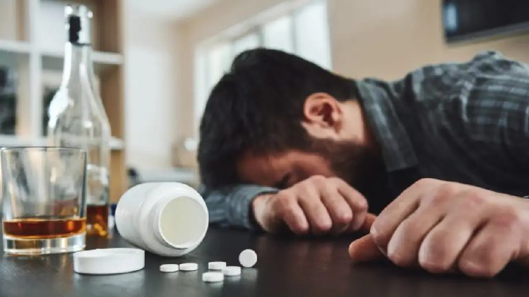 A man unconscious on a table with pills and alcohol bottle in the foreground, showing the immediate danger and need for medical drug detox rehab.