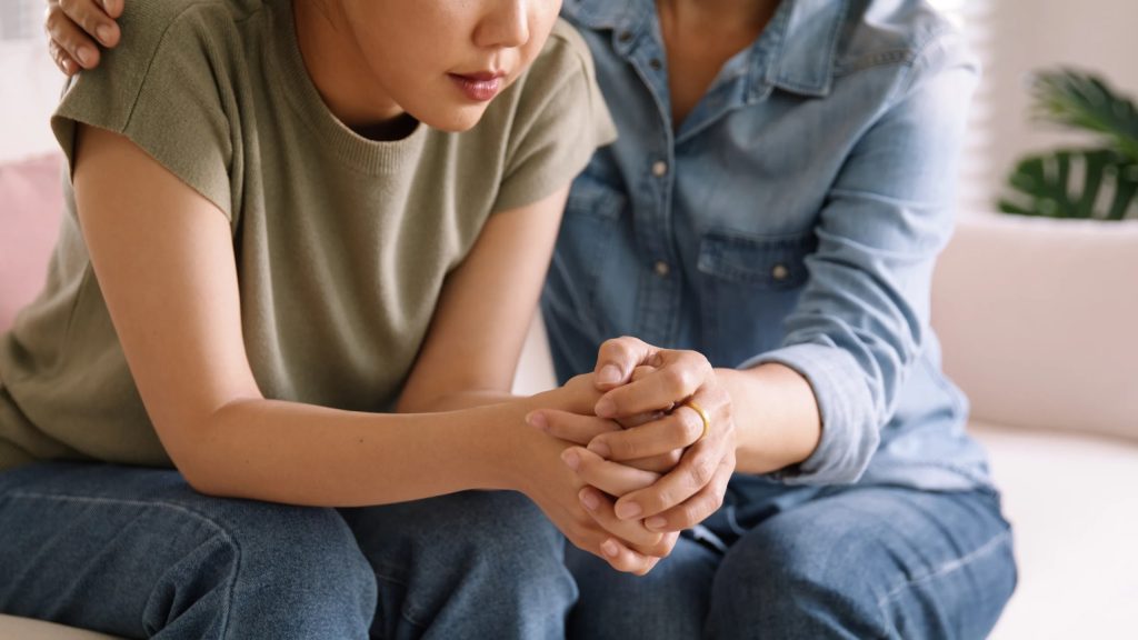 Mother holding hands and comforting her daughter on the sofa, showing family support in addiction recovery